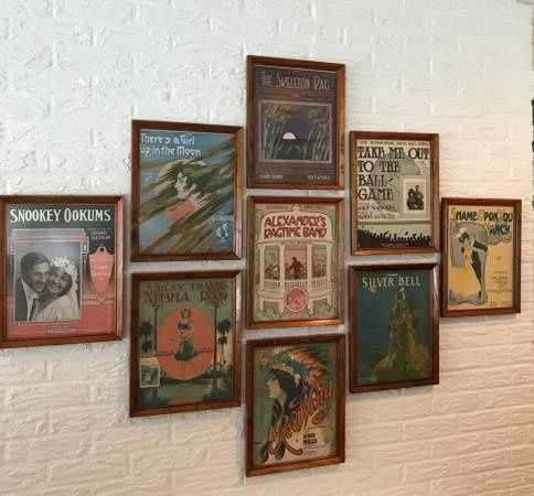 Gerahmte historische Notenblätter und Musikplakate an einer weißen Ziegelwand